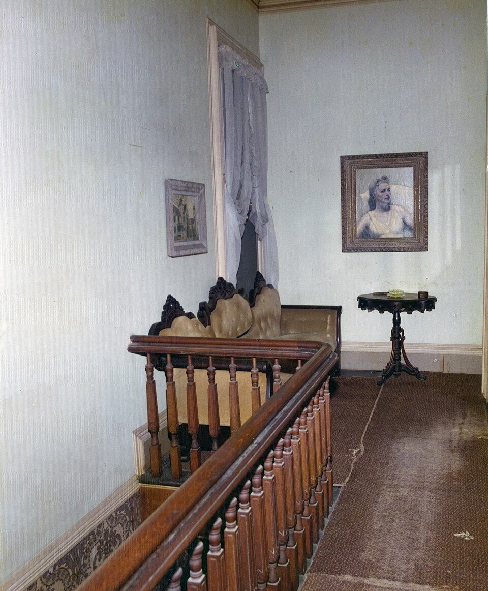 Archival view of a wooden stair railing, upholstered seat and portraits at 534 Eaton Street