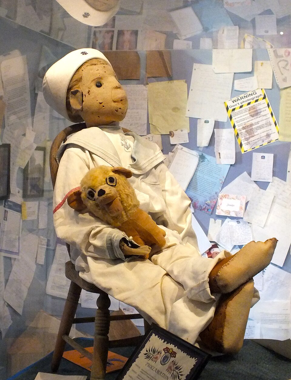Robert the Doll in a sailor suit holding a stuffed animal inside a museum case surrounded by letters